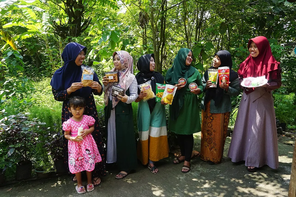 Enam perempuan berkerudung dan seorang anak perempuan berdiri di kebun sambil memegang produk camilan berkemasan, seperti keripik dalam plastik dan kue dalam kotak