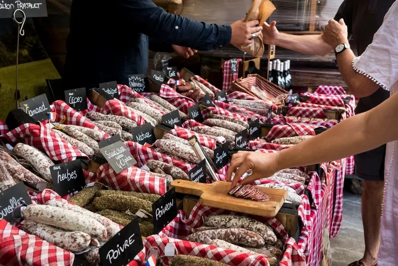 Lapak sosis kering di pasar beralas kain kotak merah putih, papan kapur kecil bertuliskan rasa seperti Poivre dan Herbes, sebuah tangan mengambil irisan sosis di atas talenan kayu sementara pedagang menyerahkan kantong kertas