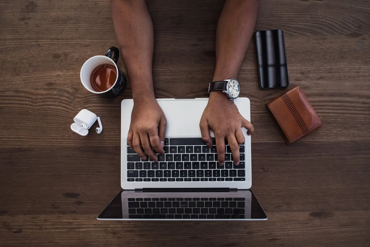 Tangan mengetik di laptop dilihat dari atas, dengan ponsel, dompet, earphone nirkabel, jam tangan, dan mug berisi minuman di meja kayu