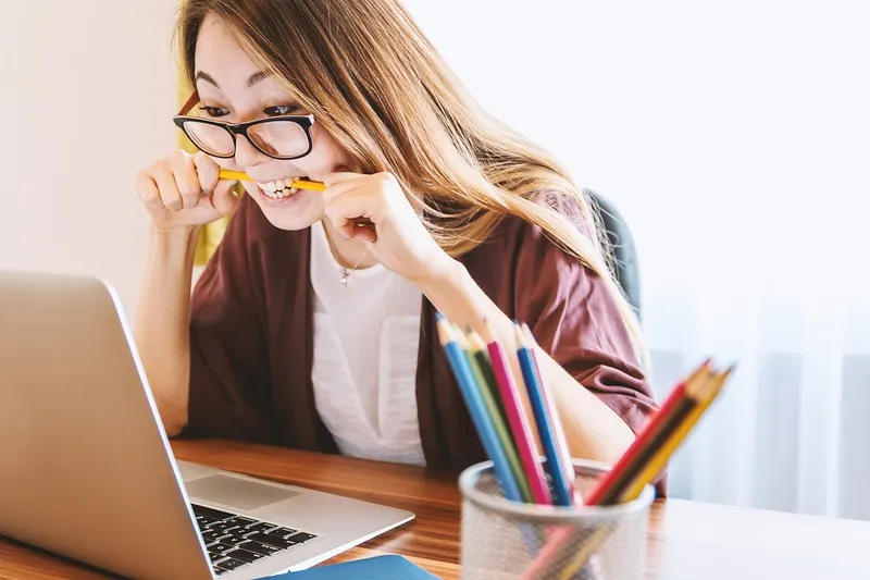 Perempuan berkacamata menggigit pensil sambil menatap laptop, dengan wadah pensil warna di meja