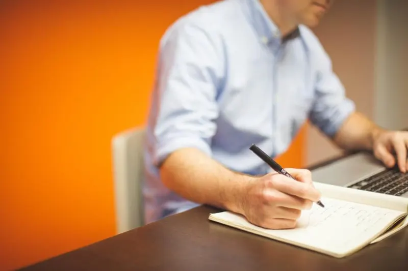 Seseorang menulis catatan di buku tulis di samping laptop yang terbuka